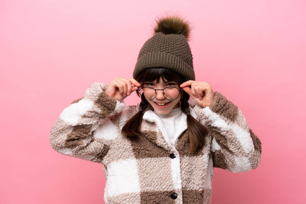 Little caucasian girl with winter jacket isolated on pink background with glasses and surprised - Foto, imagen
