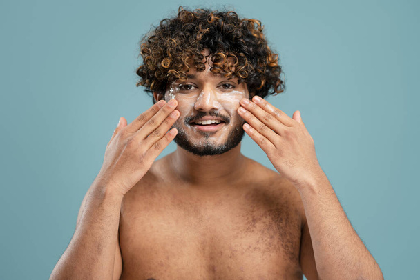 Close-up portret van jonge gekrulde bebaarde Aziatische man die een vochtinbrengende crème op zijn gezicht verspreidt, met een naakt bovenlichaam geïsoleerd over een blauwe achtergrond en glimlachend kijkend naar de camera. Huidverzorgingsconcept - Foto, afbeelding
