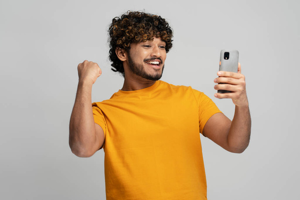 Sonriente chico asiático, estudiante en camiseta amarilla sosteniendo el teléfono móvil, apretando el puño de la emoción mientras pasa los exámenes a la universidad, obteniendo buenos resultados, expresando emociones positivas, fondo gris - Foto, Imagen