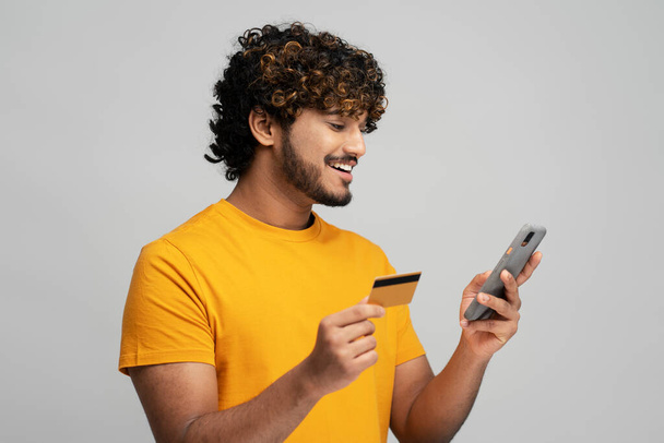 Όμορφος χαμογελαστός Ασιάτης γενειοφόρος άντρας που κρατάει πιστωτική κάρτα και χρησιμοποιεί κινητό τηλέφωνο, εισάγει δεδομένα ενώ κάνει ηλεκτρονικές συναλλαγές, μεταφέρει χρήματα μέσω internet banking, απομονωμένο γκρι φόντο - Φωτογραφία, εικόνα