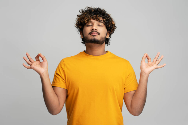 リラックスした巻き毛の若いインド人男性-明るい黄色のTシャツのヨギは、灰色の背景に彼の閉じた目で禅のポーズで瞑想します。ヒンドゥ教徒の男がヨガを。瞑想。リラックス。心の底 - 写真・画像