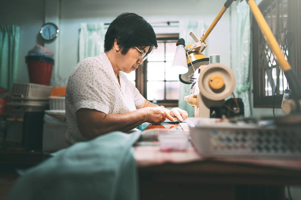 Ανάκτηση ράφτη νοτιοανατολική Ασία ηλικιωμένοι χειροτεχνία ένα πανί με ραπτομηχανή. Μωρό γενιά boomer ώριμη εργασία στο σπίτι έννοια. Στιγμές ζωής σε αυθεντικούς ανθρώπους. - Φωτογραφία, εικόνα