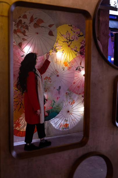 Λευκή λευκή γυναίκα με μαύρα μαλλιά. Πορτρέτο στον καθρέφτη με πολύχρωμες ιαπωνικές ομπρέλες, αστικό περιβάλλον. - Φωτογραφία, εικόνα