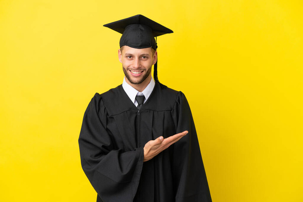 Jonge afgestudeerde blanke man geïsoleerd op gele achtergrond presenteren van een idee, terwijl hij glimlacht in de richting van - Foto, afbeelding