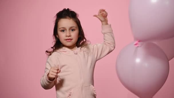 Cute Caucasian 5 years old child, a lovely baby girl is playing with inflatable helium pink pastel balloons, smiles a beautiful toothy smile looking at camera, isolated over pink color background - Imágenes, Vídeo