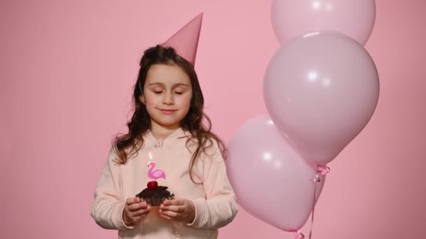Kaukasische fröhliche Kindermädchen in rosa Geburtstagskappe, die gehegten Wunsch und bläst Kerzen auf ihre Geburtstagstorte, steht in der Nähe von schönen aufblasbaren Heliumballons auf isoliertem rosa Hintergrund. - Filmmaterial, Video