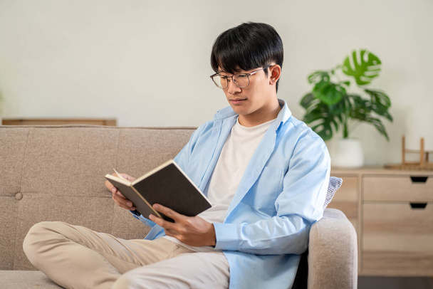 Businessman in casual is sitting on comfortable sofa to relaxation with hobby of reading the book while leisure with cozy lifestyle in living room at home. - Фото, изображение