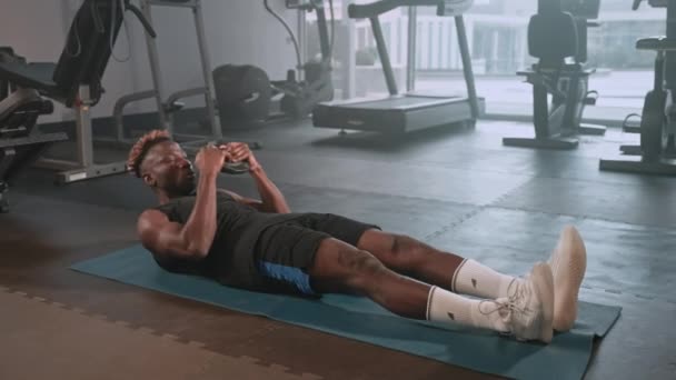 Filmagem em câmera lenta de um esportista negro fazendo sit-ups com placa de peso deitado no tapete no clube de fitness. Atleta trabalhando em abs - Filmagem, Vídeo