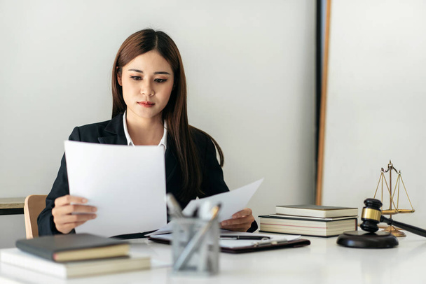 Business lawyer woman holding business contract to reading while sitting to working on the table with brass scales and justice hammer in law firm office. - Photo, Image