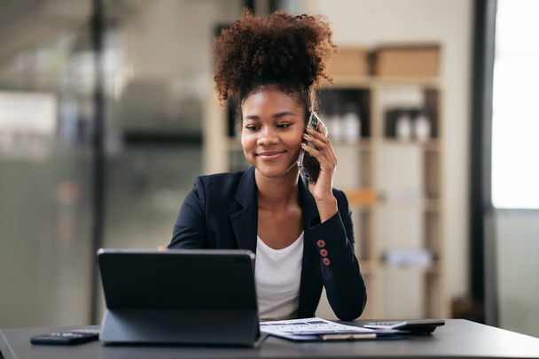 Afrikanische amerikanische Geschäftsfrau im Anzug liest Buchhaltungsinformationen über neue Geschäfte auf dem Tablet und spricht auf dem Smartphone, während sie an einem digitalen Tablet mit Papierkram am modernen Arbeitsplatz arbeitet. - Foto, Bild