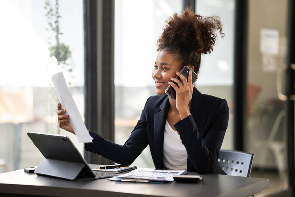 African american businesswoman in suit holding paperwork to reading information and talking on smartphone while working about accounting of new business on digital tablet in modern workspace. - Foto, Bild