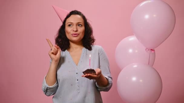 Delightful woman in festive cap, enumerating her wishes, reasoning on plans, counting on fingers, celebrating birthday party, blowing candle on cupcake, smiling looking at camera on a pink background - Filmati, video