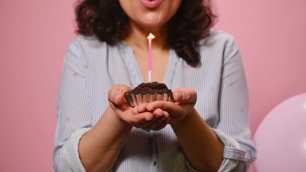 Selective focus on a delicious sweet chocolate cupcake in the hands of smiling birthday girl, blowing candle on pink color background with inflatable helium balloons. Woman celebrating birthday party - Filmati, video