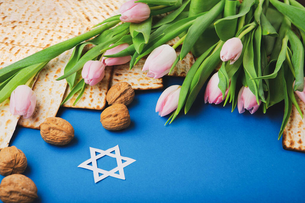 Jewish holiday Passover greeting card concept with matzah, nuts, tulip flowers on blue table Seder Pesach spring holiday background, top view, copy space. - Fotó, kép
