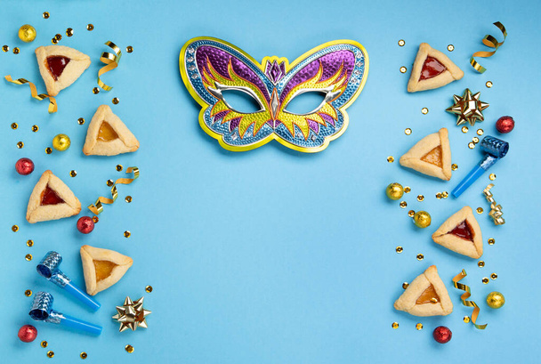 Purim celebration jewish carnival holiday concept. Tasty hamantaschen cookies, Carnival mask, noisemaker, sweet candies and party decor on blue background. Top view, flat lay, copy space. - Photo, Image