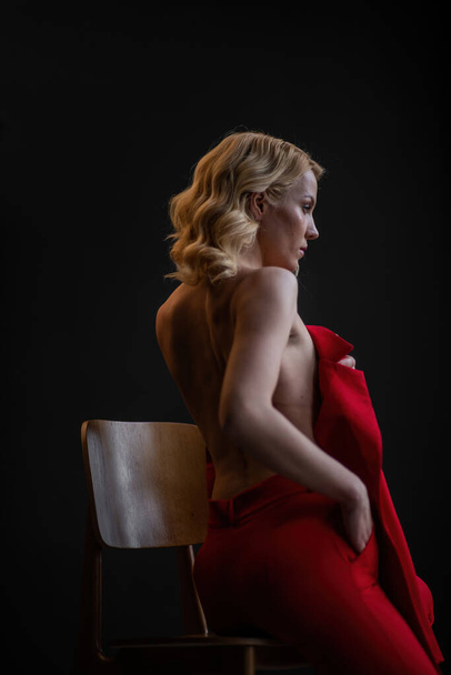 Adult beauty woman bare back in formal evening red trousers sitting pose without bra. Stylish blonde curly hair sensual nude model fashionista posing at studio in fashion pantsuit out of blazer - Zdjęcie, obraz