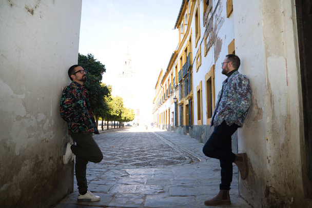 Young couple of gay men, they are leaning on an arch and in the background you can see the cathedral of seville and the orange trees in the garden. The couple is married. Concept of gay rights - 写真・画像