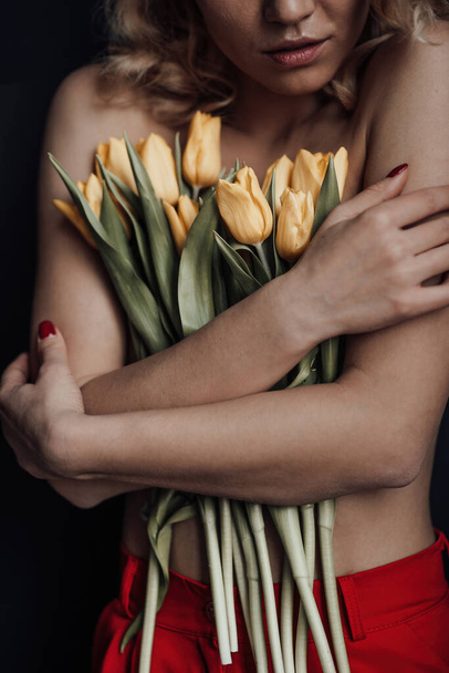 Dorosła piękna kobieta pół naga w formalnych czerwonych spodniach wieczorowych bez biustonosza przytula bukiet żółtych tulipanów. stylowa blondynka kręcone włosy zmysłowy nagie model mody pozowanie w studio w wiosenne wakacje - Zdjęcie, obraz