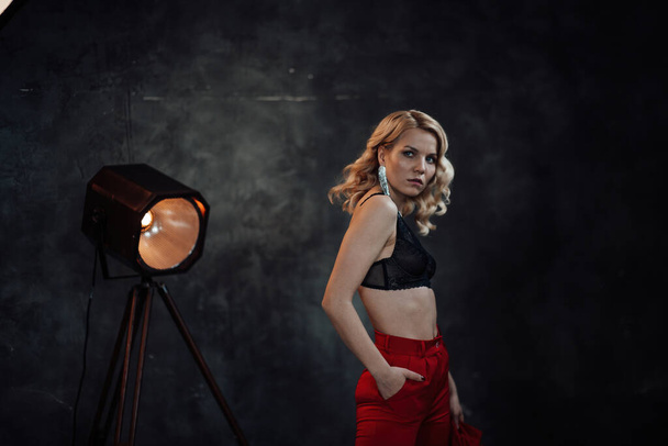 Young adult beauty woman in formal evening suit of red color with lace black bra underwear standing by thoughtful. Stylish blonde curly hair model fashionista posing at studio in fashion pantsuit - Foto, Imagen