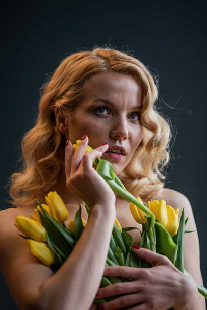 Adult beauty woman half naked in formal evening red trousers without bra hugs bouquet of yellow tulips. Stylish blonde curly hair sensual nude model fashionista posing in studio at spring holidays - Fotografie, Obrázek