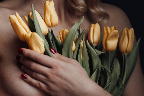 Felnőtt szépség nő félmeztelen formális este piros nadrág melltartó nélkül ölelés csokor sárga tulipán. Stílusos szőke göndör haj érzéki meztelen modell fashionista pózol a stúdióban a tavaszi szünetben - Fotó, kép