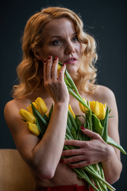Dospělá krasavice polonahá ve večerních červených kalhotách bez podprsenky objímá kytici žlutých tulipánů. Stylová blondýnka kudrnaté vlasy smyslný nahota model fashionista pózování ve studiu na jarní prázdniny - Fotografie, Obrázek