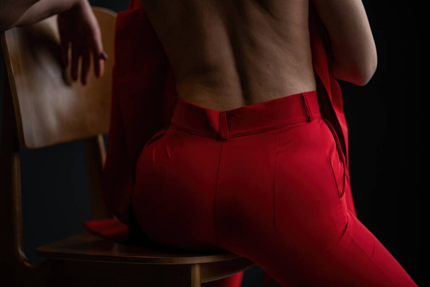 Mujer de belleza adulta desnuda de nuevo en pantalones rojos de noche formal sentado pose sin sujetador. Elegante rubia pelo rizado sensual modelo desnudo fashionista posando en el estudio en traje pantalón de moda fuera de chaqueta - Foto, imagen