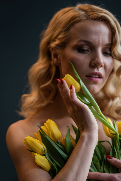 Adult beauty woman half naked in formal evening red trousers without bra hugs bouquet of yellow tulips. Stylish blonde curly hair sensual nude model fashionista posing in studio at spring holidays - Zdjęcie, obraz