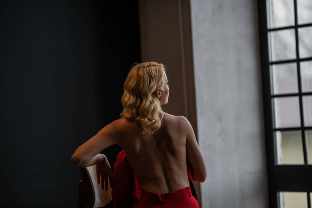 Volwassen schoonheid vrouw naakt terug in formele avond rode broek zitten pose zonder beha. Stijlvol blond krullend haar sensueel naakt model fashionista poseren in studio in mode broekpak uit blazer - Foto, afbeelding