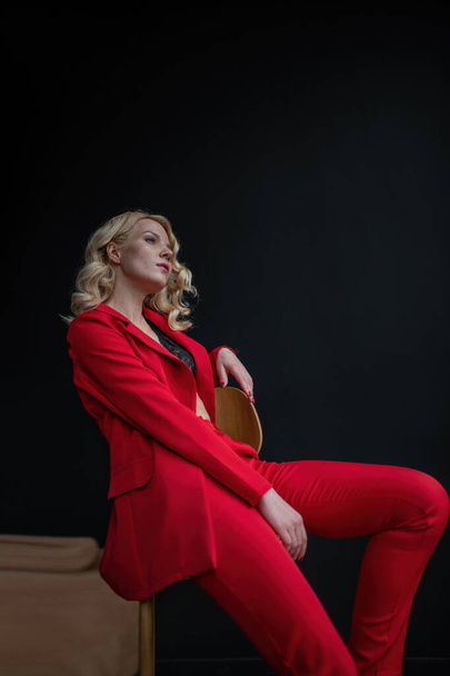 Young adult beauty woman in formal evening suit of red color with lace black bra underwear standing by thoughtful. Stylish blonde curly hair model fashionista posing at studio in fashion pantsuit - 写真・画像