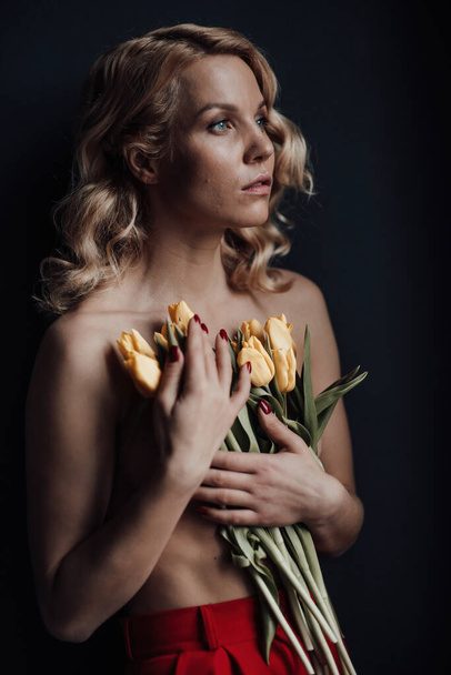 Adult beauty woman half naked in formal evening red trousers without bra hugs bouquet of yellow tulips. Stylish blonde curly hair sensual nude model fashionista posing in studio at spring holidays - Photo, image
