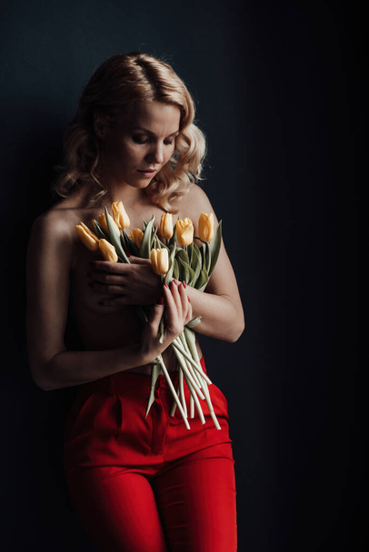 Adult beauty woman half naked in formal evening red trousers without bra hugs bouquet of yellow tulips. Stylish blonde curly hair sensual nude model fashionista posing in studio at spring holidays - Photo, image