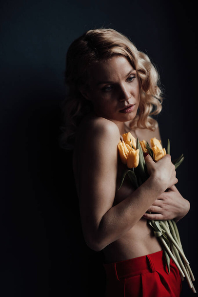 Adult beauty woman half naked in formal evening red trousers without bra hugs bouquet of yellow tulips. Stylish blonde curly hair sensual nude model fashionista posing in studio at spring holidays - Zdjęcie, obraz
