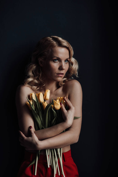 Adult beauty woman half naked in formal evening red trousers without bra hugs bouquet of yellow tulips. Stylish blonde curly hair sensual nude model fashionista posing in studio at spring holidays - Foto, Imagen
