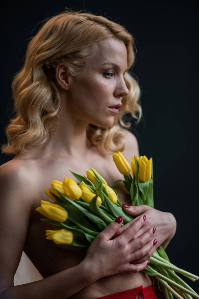 Adult beauty woman half naked in formal evening red trousers without bra hugs bouquet of yellow tulips. Stylish blonde curly hair sensual nude model fashionista posing in studio at spring holidays - Photo, Image