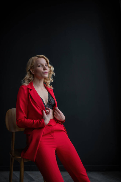 Young adult beauty woman in formal evening suit of red color with lace black bra underwear standing by thoughtful. Stylish blonde curly hair model fashionista posing at studio in fashion pantsuit - Fotografie, Obrázek