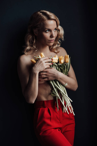 Adult beauty woman half naked in formal evening red trousers without bra hugs bouquet of yellow tulips. Stylish blonde curly hair sensual nude model fashionista posing in studio at spring holidays - Fotó, kép