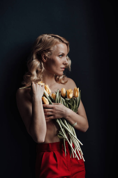 Adult beauty woman half naked in formal evening red trousers without bra hugs bouquet of yellow tulips. Stylish blonde curly hair sensual nude model fashionista posing in studio at spring holidays - Foto, Imagen