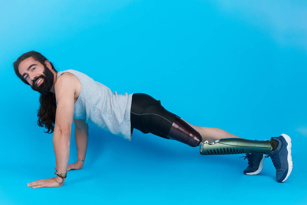 Horizontales Studioporträt mit blauem Hintergrund eines glücklichen Mannes mit Beinprothese beim Liegestütz - Foto, Bild