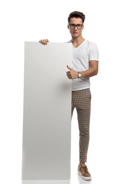 lässiger junger Mann zeigt weiße Plakatwand und macht Daumen nach oben auf weißem Hintergrund  - Foto, Bild
