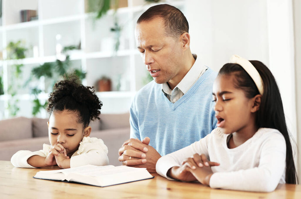 Family, worship and bible with father and kids praying at table for peace, religion and faith in their home. God, pray and children with parent for prayer, bible study or Jesus Christ praise together. - Photo, Image