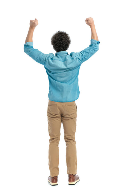 back view of curly hair guy holding arms above head and cheering victory in front of white background in studio - Valokuva, kuva