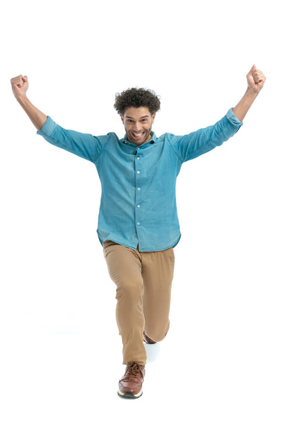 full body picture of enthusiastic young man with arms above head bending the knee and celebrating in front of white background in studio - Photo, Image