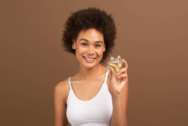 Smiling young black curly lady with perfect skin hold bottle of perfume, recommends ad and offer isolated on brown background, studio. Cosmetics, beauty care and favorite smell, sale and fashion - Fotó, kép