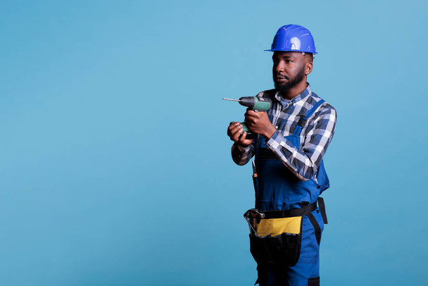 Концентрированный африканский американский строитель держит беспроводную дрель на синем фоне. Профессиональный человек с ремнем строительные инструменты носить каску и покрывало, контрататор работы. - Фото, изображение