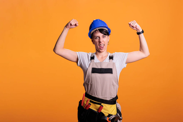Confident female contractor flexing arm muscles, preparing to work on renovation project with tools. Confident energetic woman pointing biceps and triceps, studio shot on yellow background. - Foto, Imagen