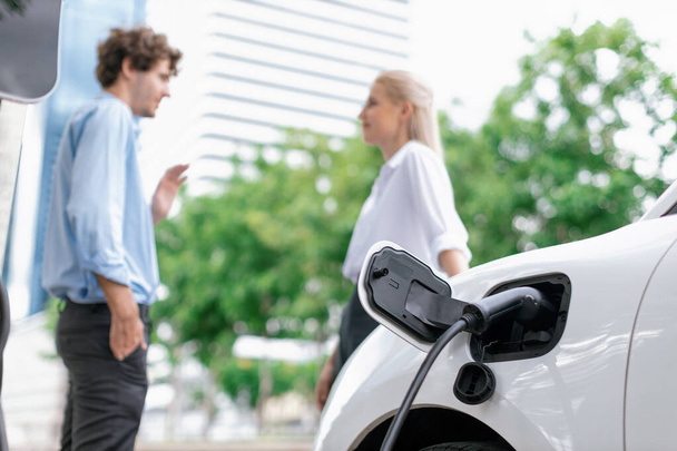 Focus parking-electric car connected to public charging station with blur progressive businesspeople holding coffee, residential building apartment and condo background for eco-friendly concept. - Photo, image