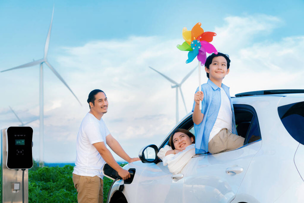 Concept of progressive happy family holding windmill toy and relax at wind farm with electric vehicle. Electric vehicle driven by clean renewable energy from wind turbine generator to charger station. - Foto, immagini