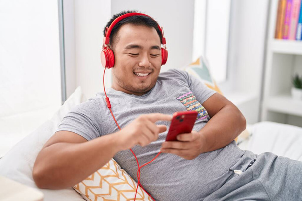 Young chinese man listening to music lying on bed at bedroom - Fotó, kép