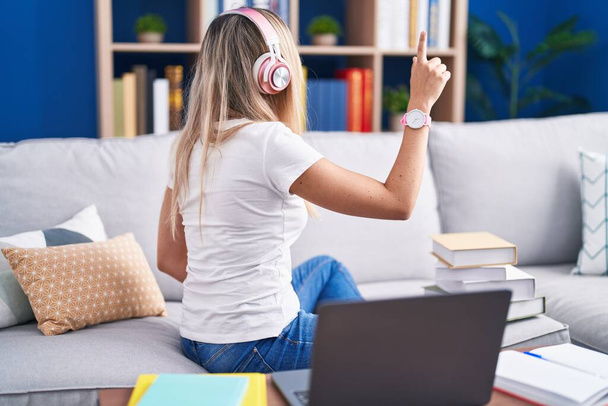 Jeune femme blonde étudiant à l'aide d'un ordinateur portable à la maison posant vers l'arrière pointant vers l'avant avec la main du doigt  - Photo, image